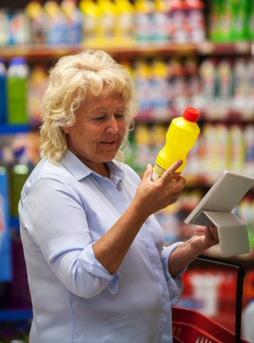 Senior woman choosing detergent in the shop. She checking the bottle label and using tablet computer to be sure with safety of goods